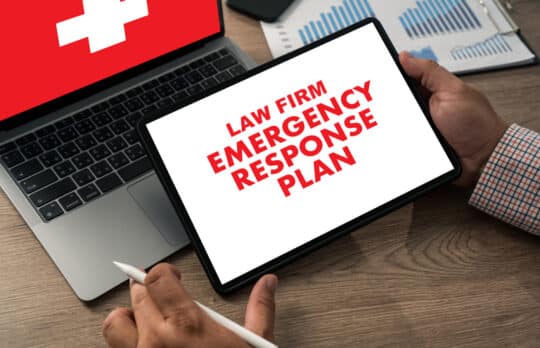 A person holding a tablet displaying a law firm emergency plan next to a laptop and charts.