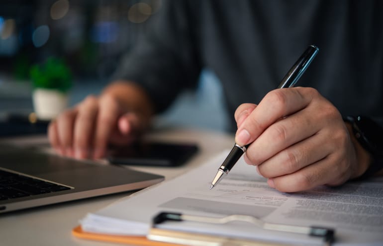 Close-up of a lawyer holding a pen to edit documents on a clipboard next to a laptop, representing legal writing.