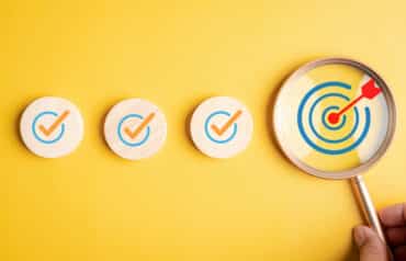 Magnifying glass focusing on a bullseye target next to checked wooden blocks, representing a comprehensive law firm startup checklist.
